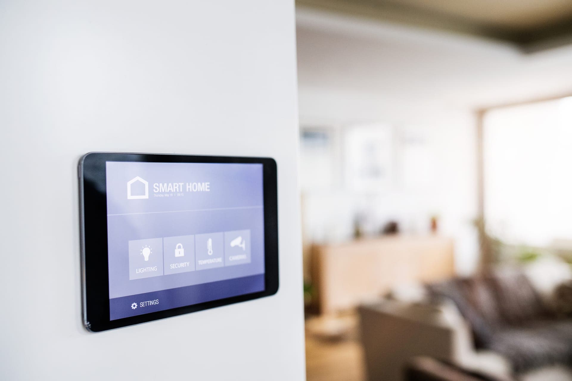 Wall-mounted smart home touchscreen control panel with a living room in the background